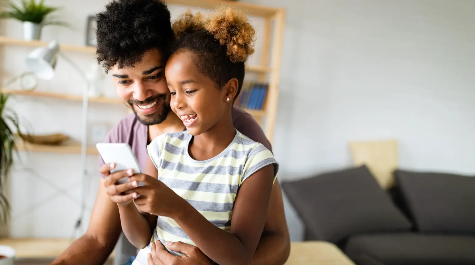 Smiling father and daughter sitting together at home looking at a smartphone.