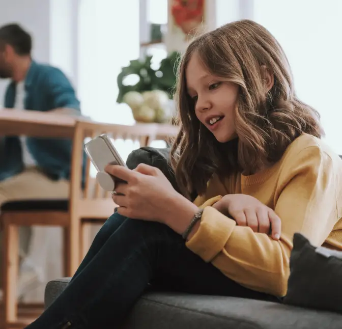 Smiling young girl sitting on a couch using a smartphone, with an adult blurred in the background at a table.