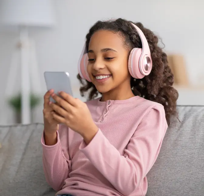 Smiling girl wearing pink headphones sitting on a couch and looking at a smartphone.