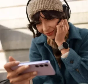 Woman wearing headphones and a beanie, smiling while watching something on her smartphone outdoors.